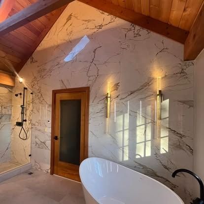 Modern bathroom with a white freestanding tub, marble-tiled wall, warm wood ceiling, and two sleek wall-mounted lights.