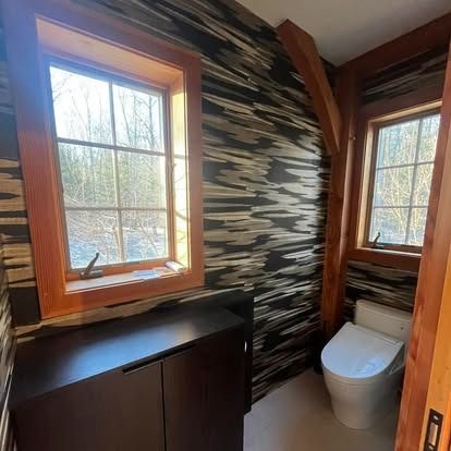 A small bathroom with bold, dark horizontal-striped wallpaper, wood-trimmed windows, a dark vanity, and a white toilet.
