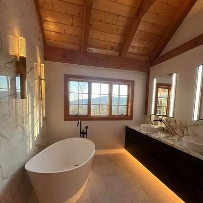 A modern bathroom with a freestanding white tub, double vanity, marble countertops, and warm wooden ceiling beams.