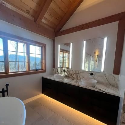 Modern bathroom with a double vanity, marble countertop, LED mirrors, and wooden ceiling beams overlooking a forest.