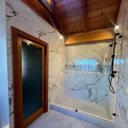 A modern bathroom with marble walls, a glass walk-in shower, and a wooden vaulted ceiling over a dark-framed door.
