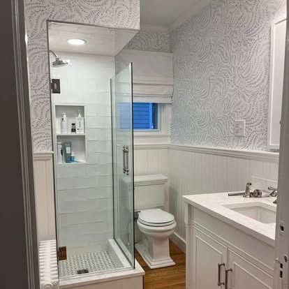 Small bathroom featuring a glass-enclosed shower, toilet, white vanity, and light blue patterned wallpaper.