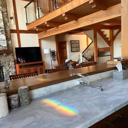 A rainbow prism reflection illuminates a gray kitchen countertop in a large, open-concept living space with wood beams.