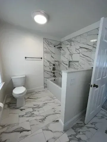 A modern bathroom with white and gray marble floors, a white toilet, and a tiled bathtub and shower alcove.