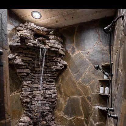 A rustic stone shower featuring a waterfall showerhead integrated into a stacked stone wall with built-in shelves.