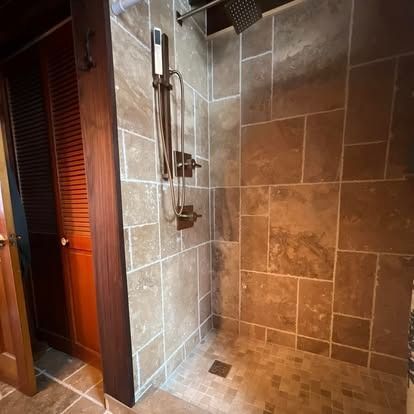 A walk-in shower with brown stone tile walls and flooring, featuring a bronze showerhead, handheld sprayer, and controls.