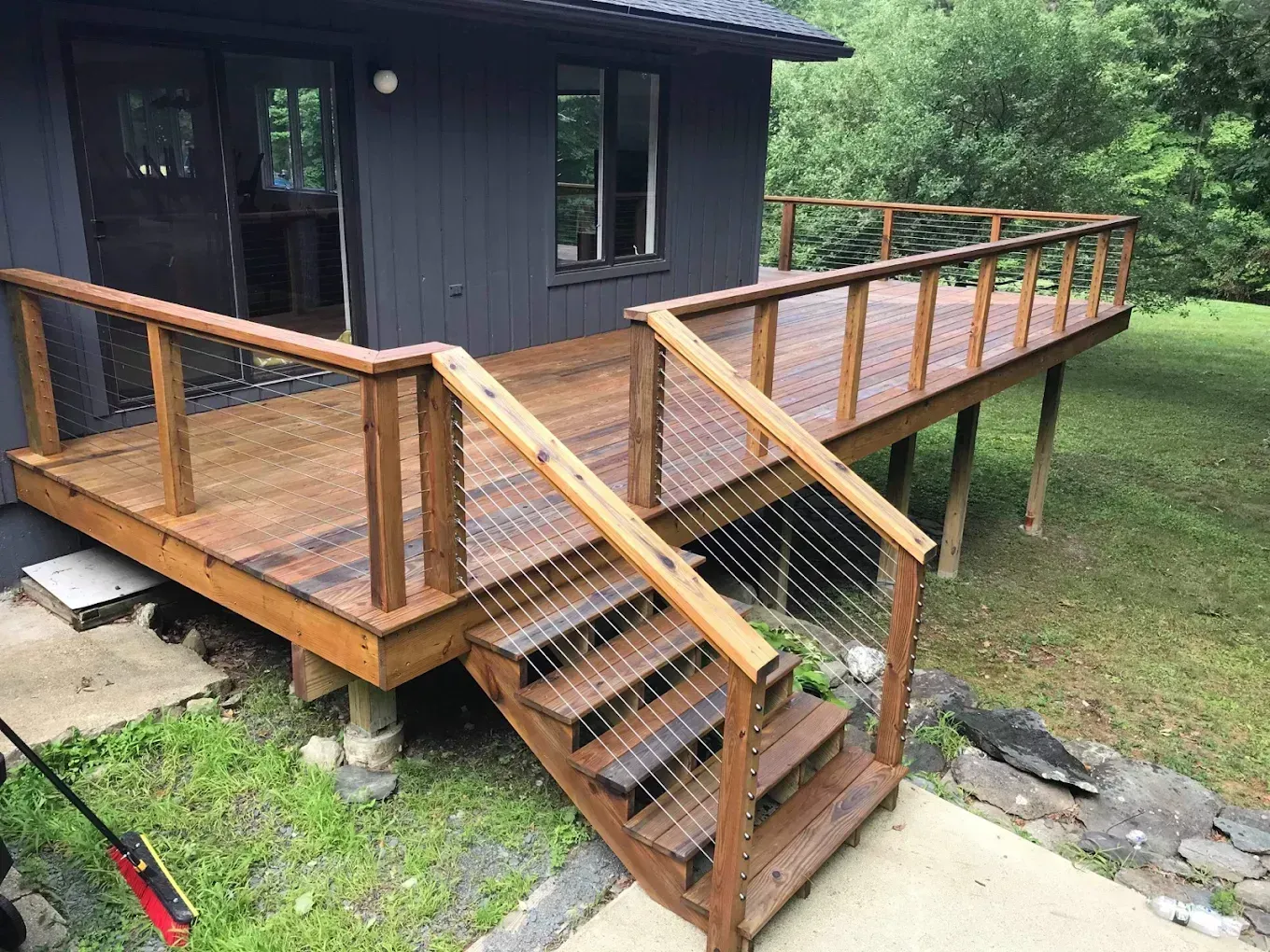 A wooden deck with cable railings and stairs, attached to a dark-sided house, overlooking a grassy yard.