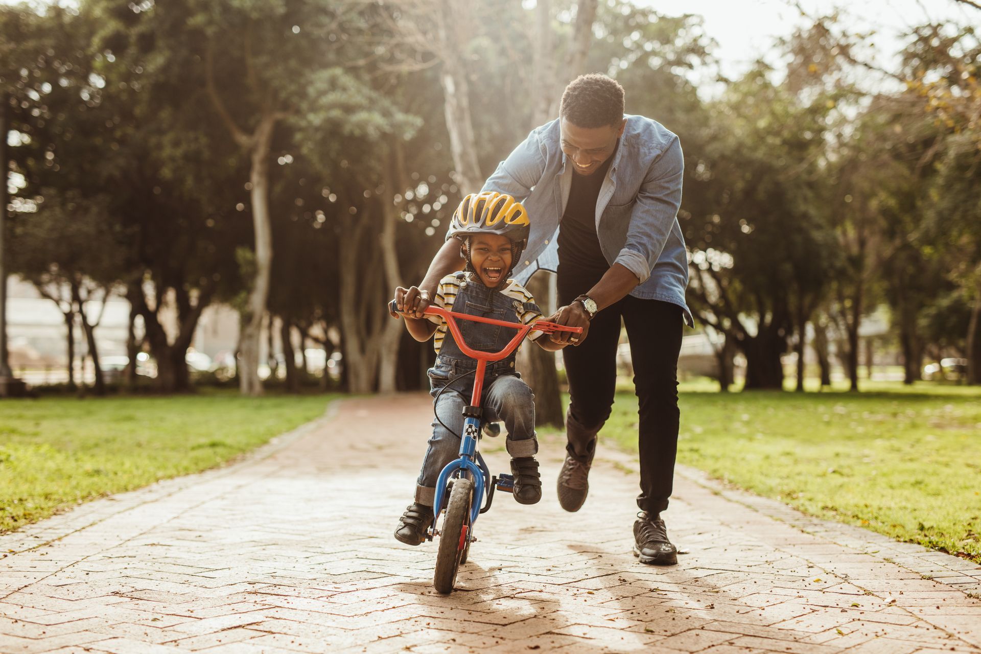 Father helping his child learn to ride a bike in a park; sunlit, happy moment.
