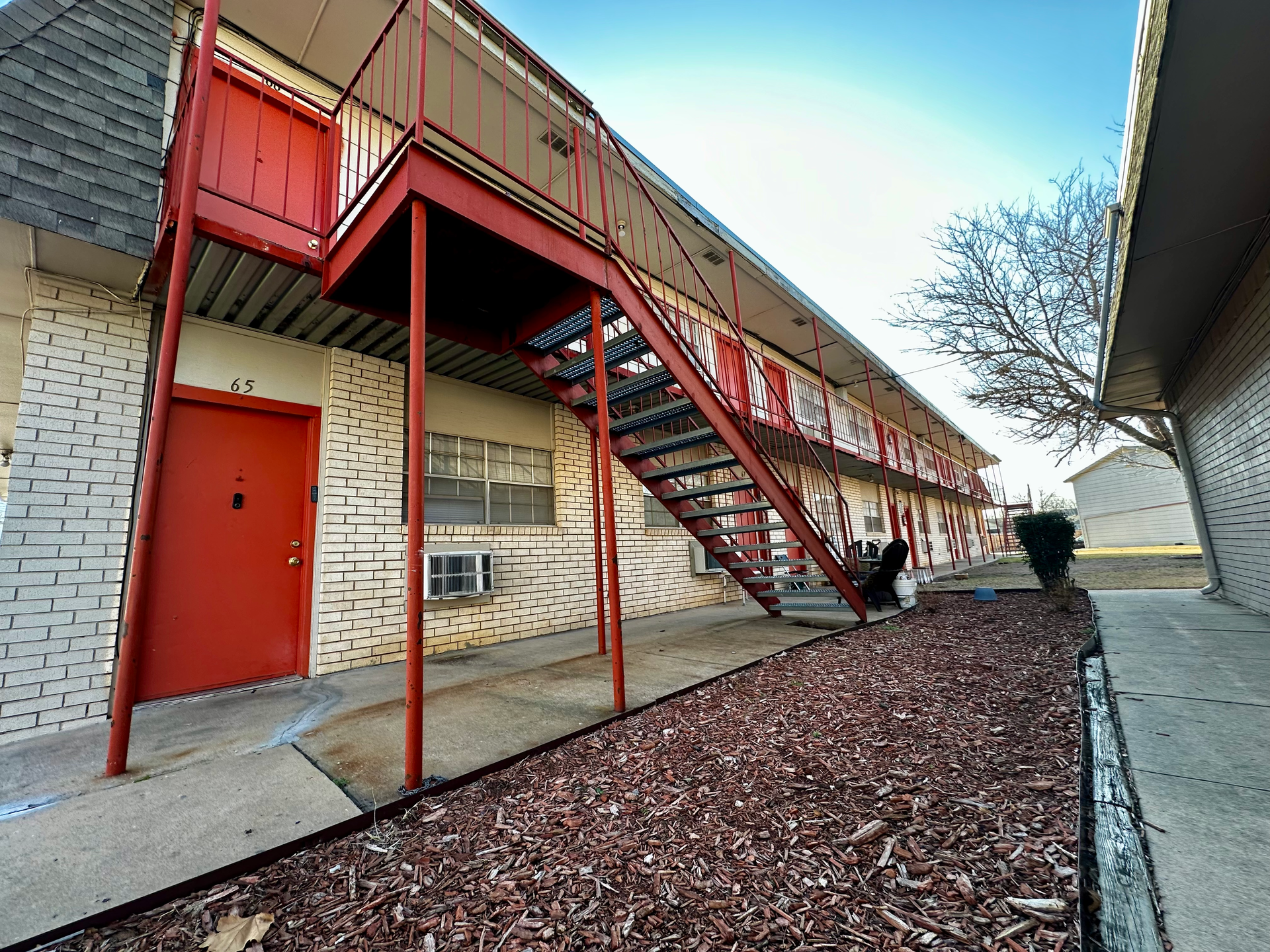 Apartment building exterior with red stairwell. Cream brick, red doors, and brown mulch. Blue sky.