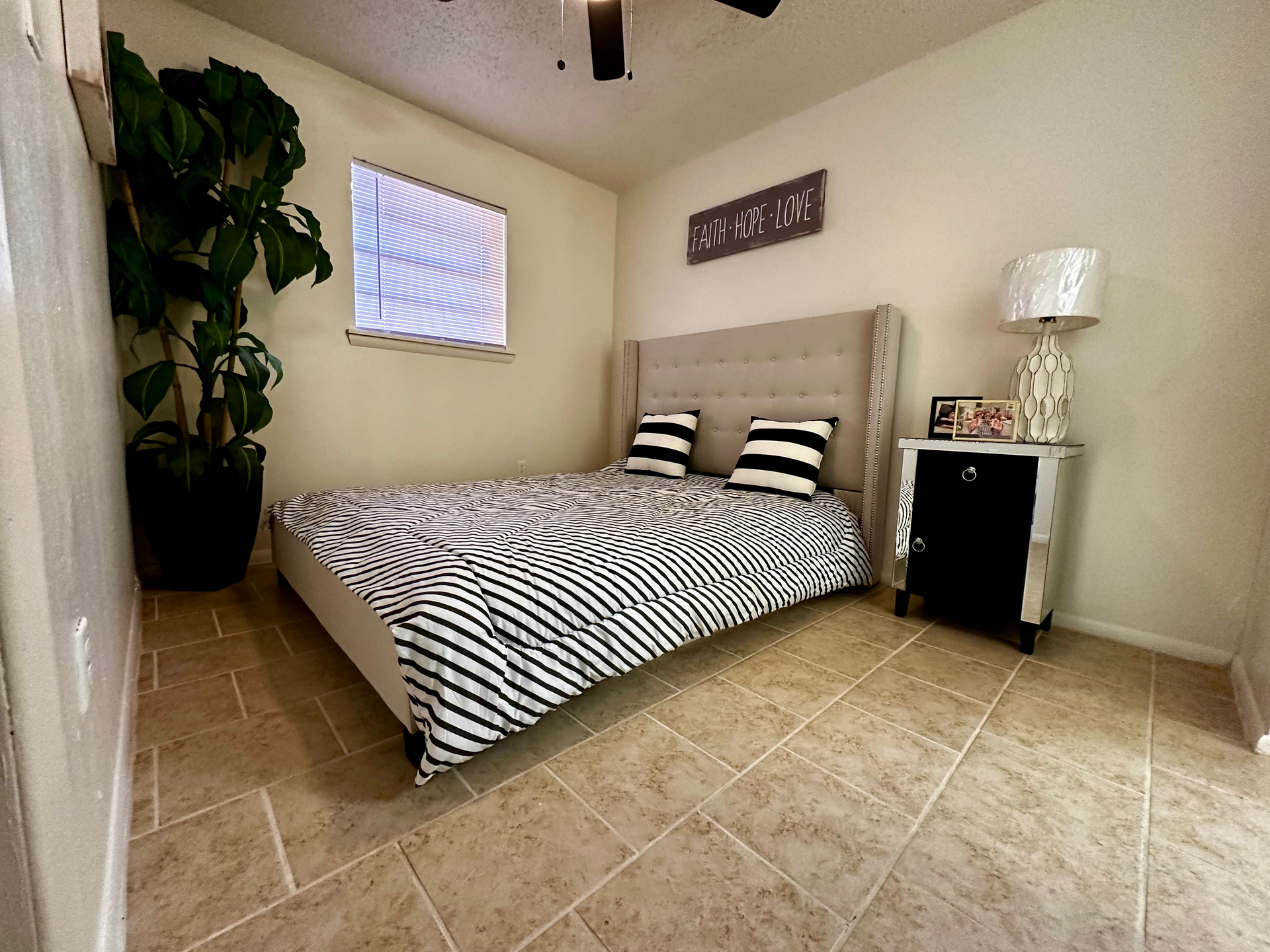 A bedroom with a bed, nightstand, potted plant, and a window. Beige walls and tile floors.
