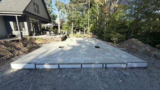 Dalle de béton autour d'une maison, terrain gravillonné, entouré de terre et d'arbres.