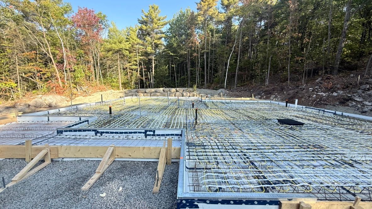 Chantier de construction avec fondations en béton préparées, treillis d'armature, coffrages en bois et arbres environnants.
