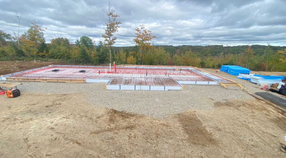 Chantier de fondations avec armatures et tuyauterie. Gravier, ciel nuageux, arbres en arrière-plan.