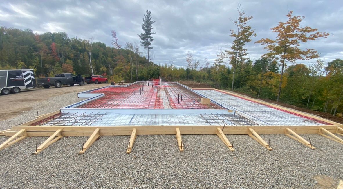 Chantier : coffrages en bois, base de gravier, treillis d'armature, tubes de chauffage radiant rouges, ciel gris.