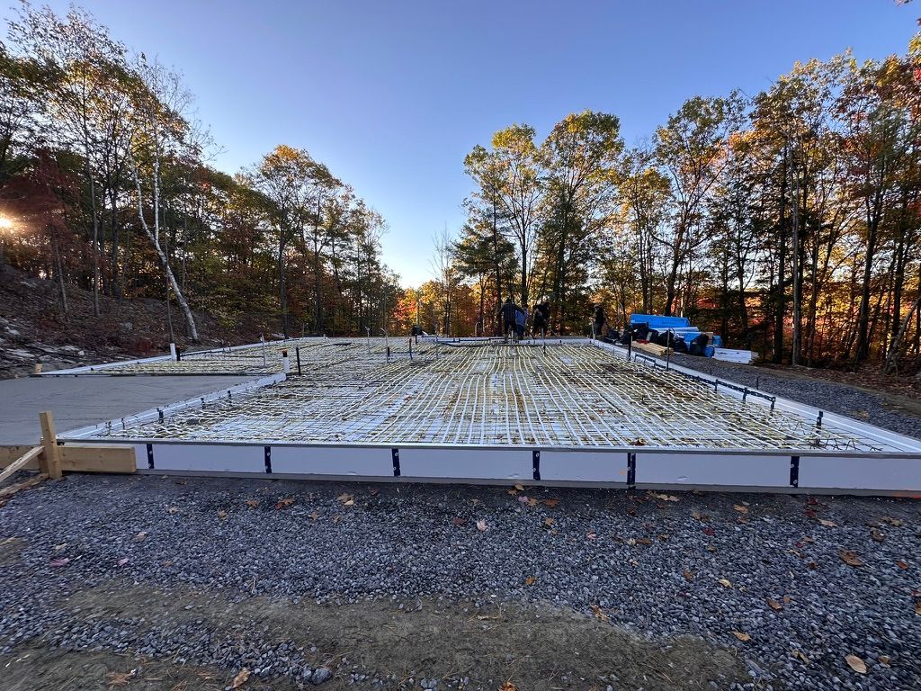 Chantier de construction de fondations avec coffrages en mousse, barres d'armature, gravier et arbres sous un ciel bleu.