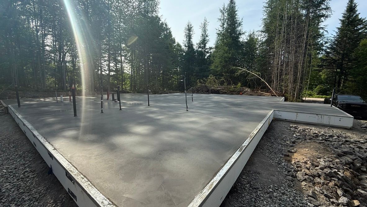 Fondation en béton fraîchement coulé dans une zone boisée ; surface grise, bordures blanches, arbres en arrière-plan.