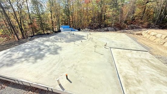 Fondations en béton fraîchement coulées avec canalisations, entourées d'un terrain vague et d'arbres.
