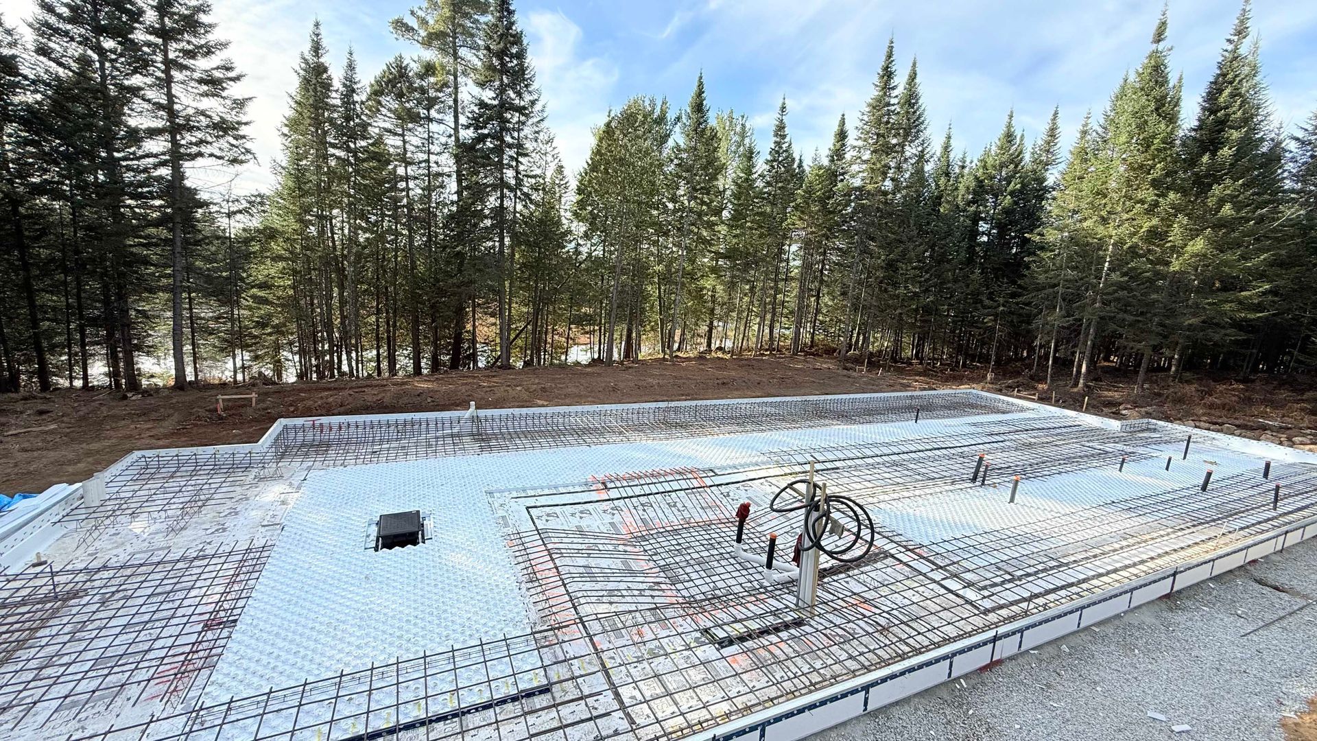 Chantier de construction avec fondations en béton et armature en acier, entouré d'arbres.