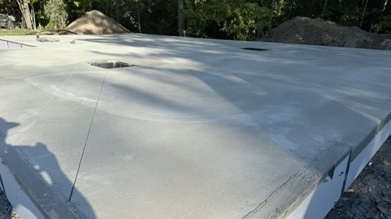 Fondation en dalle de béton gris fraîchement coulée, présentant des fissures récentes ; environnement extérieur.