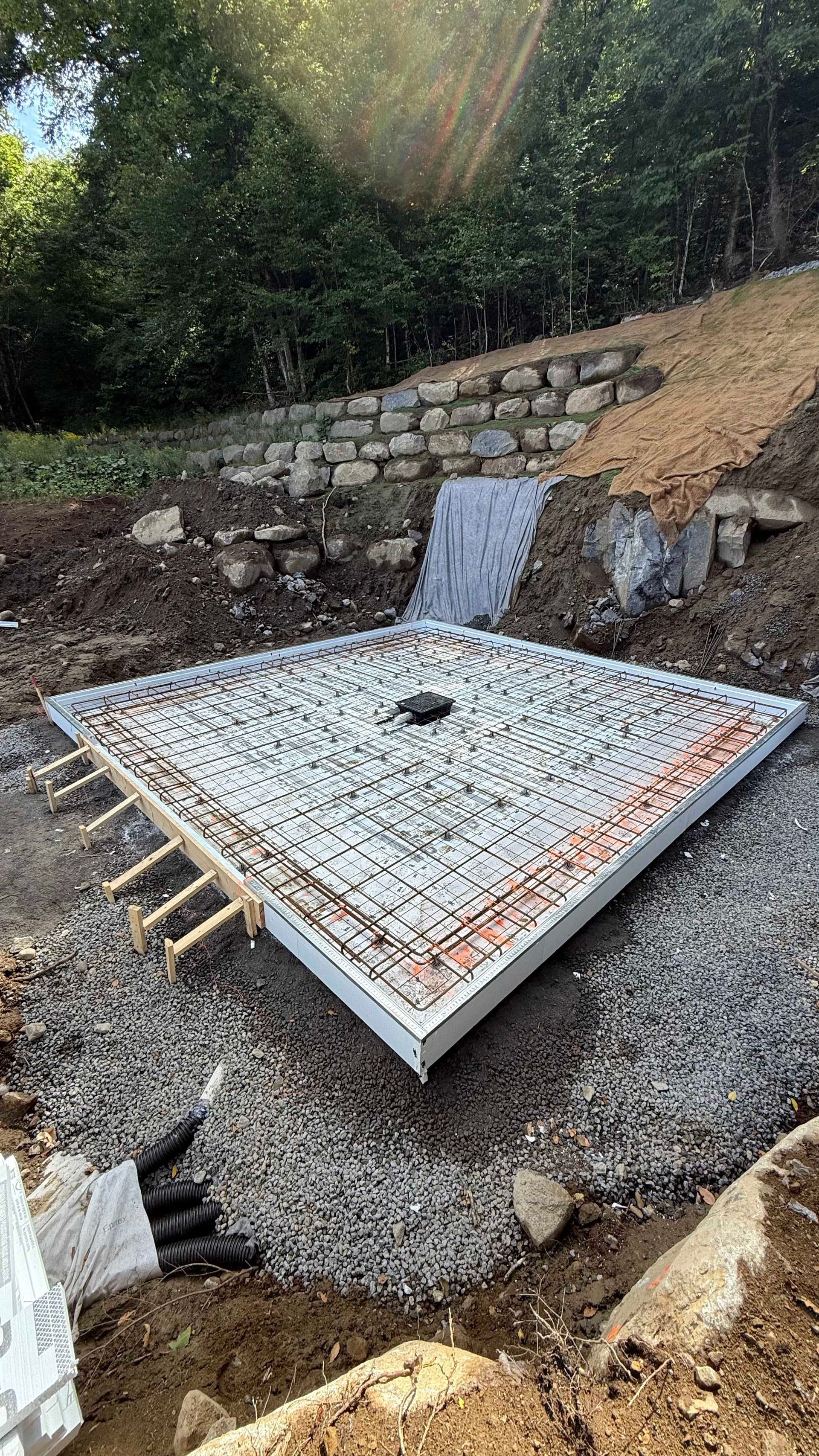 Chantier : Fondation carrée en béton entourée de gravier, flanc de colline en arrière-plan.