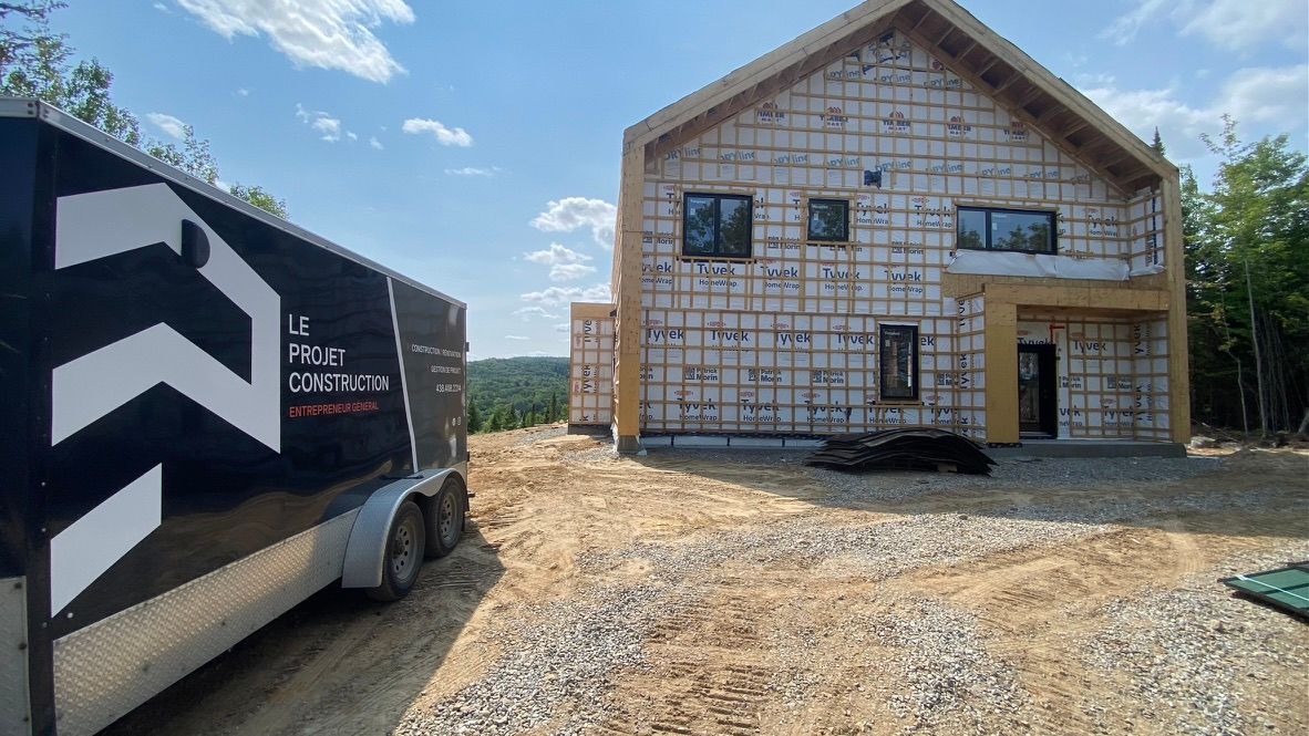 Maison en construction avec une remorque noire garée au premier plan.
