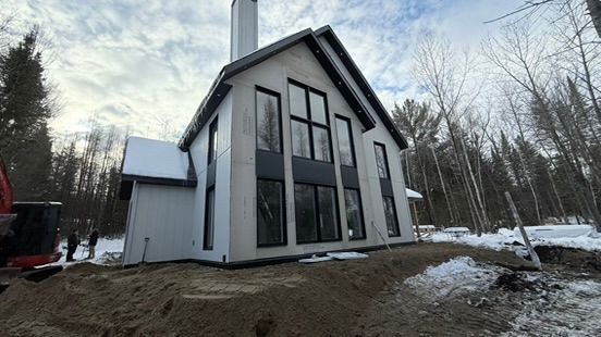 Maison blanche moderne avec de hautes fenêtres, des boiseries noires et une cheminée, au milieu d'une zone boisée enneigée.