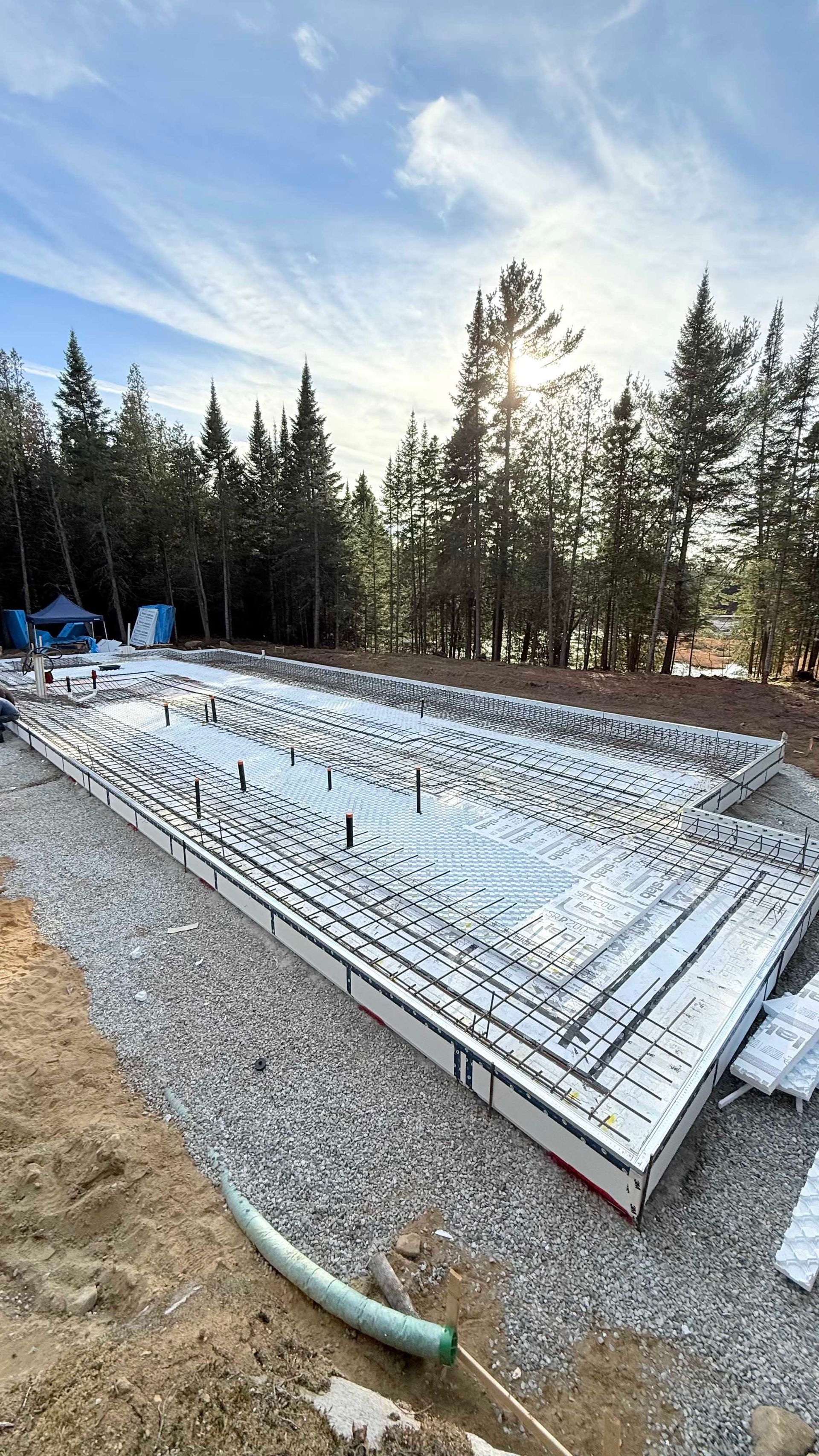 Chantier de construction avec coffrages en béton isolés, base de gravier et arbres. Ciel bleu.