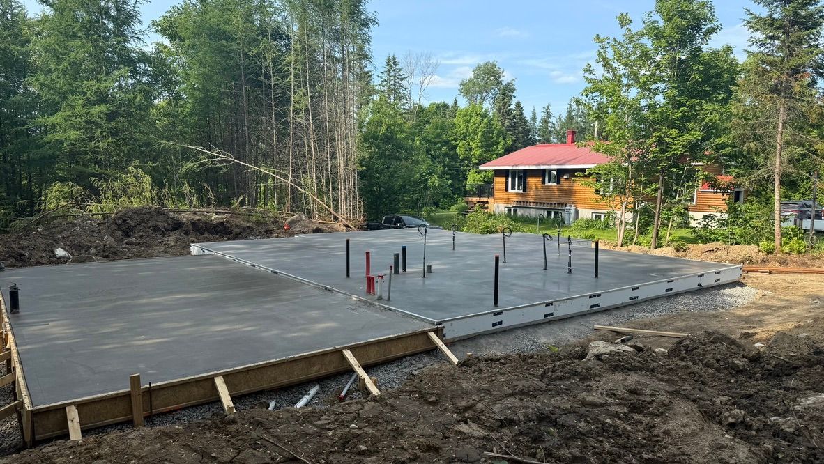Fondations en béton fraîchement coulées avec plomberie, entourées d'une zone boisée.