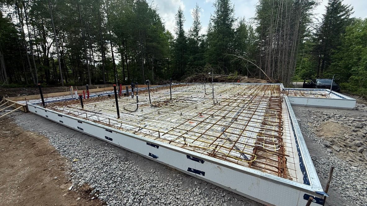 Fondations d'un bâtiment en construction, avec coffrages en béton isolés, armatures et canalisations visibles.