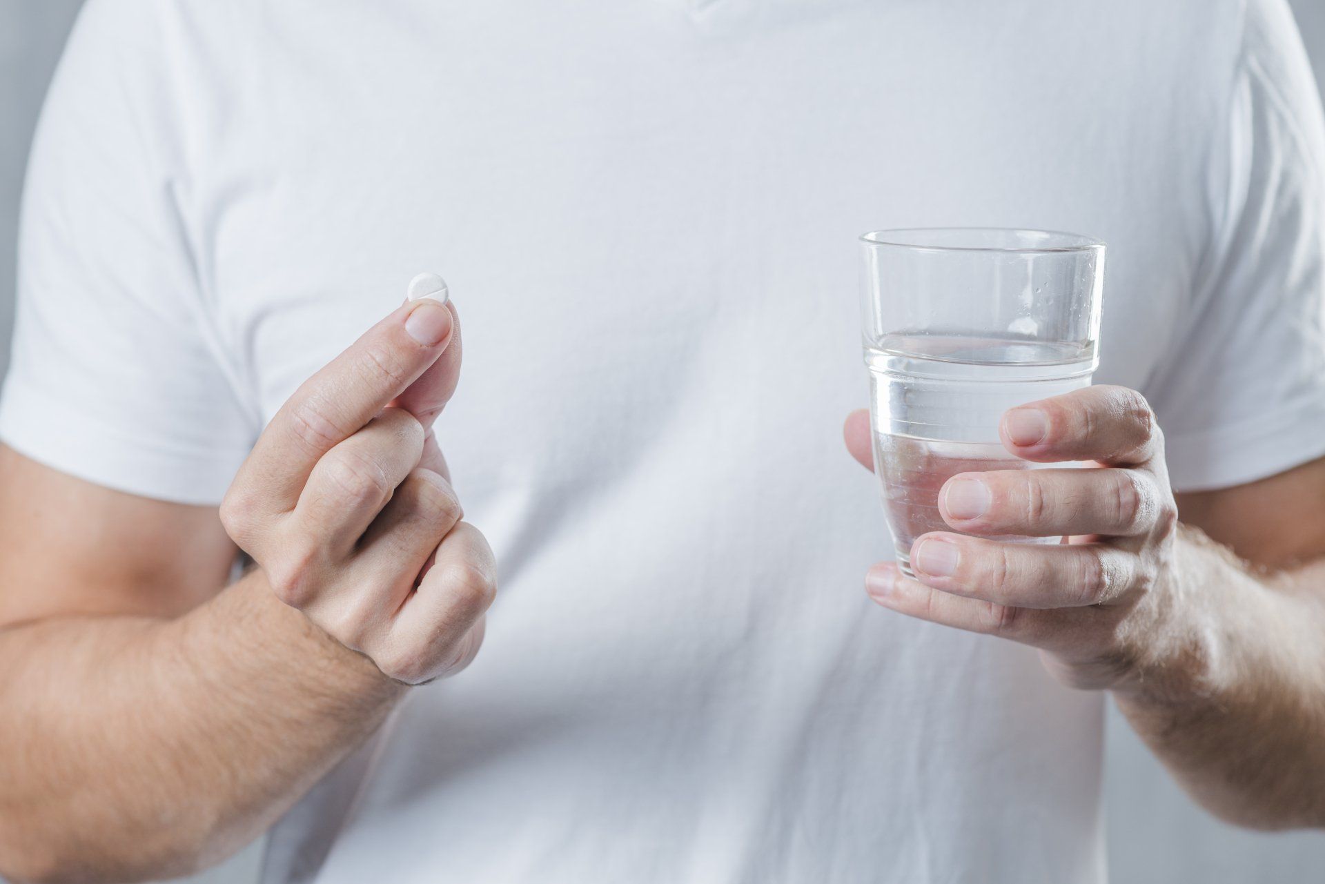 Homem vestindo camiseta branca, segurando um copo com água e um comprimido de nimesulida