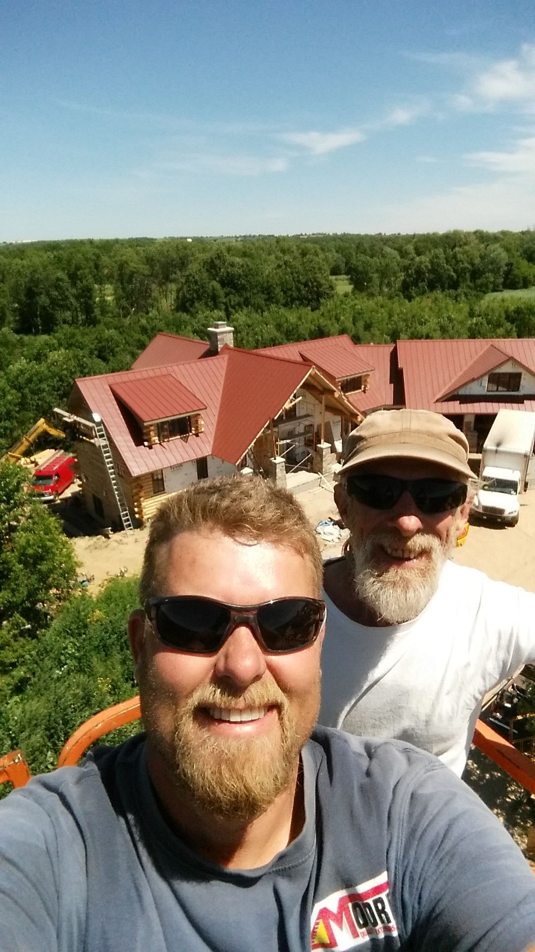 Two smiling people wearing sunglasses take a selfie high above a house under construction with a red metal roof.
