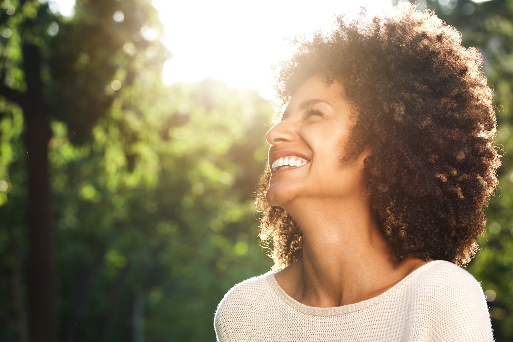A woman with curly hair is smiling in front of a forest.