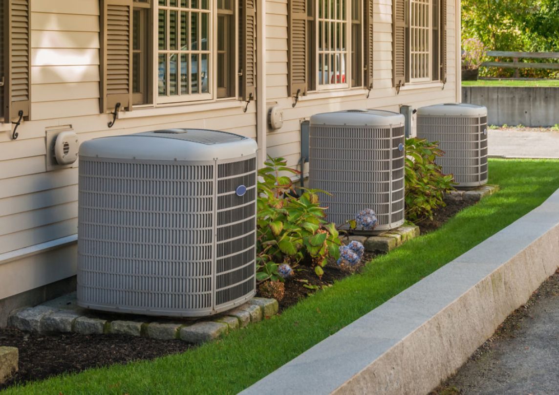 Three air conditioning units outside a white house with brown shutters, set in a small garden bed.