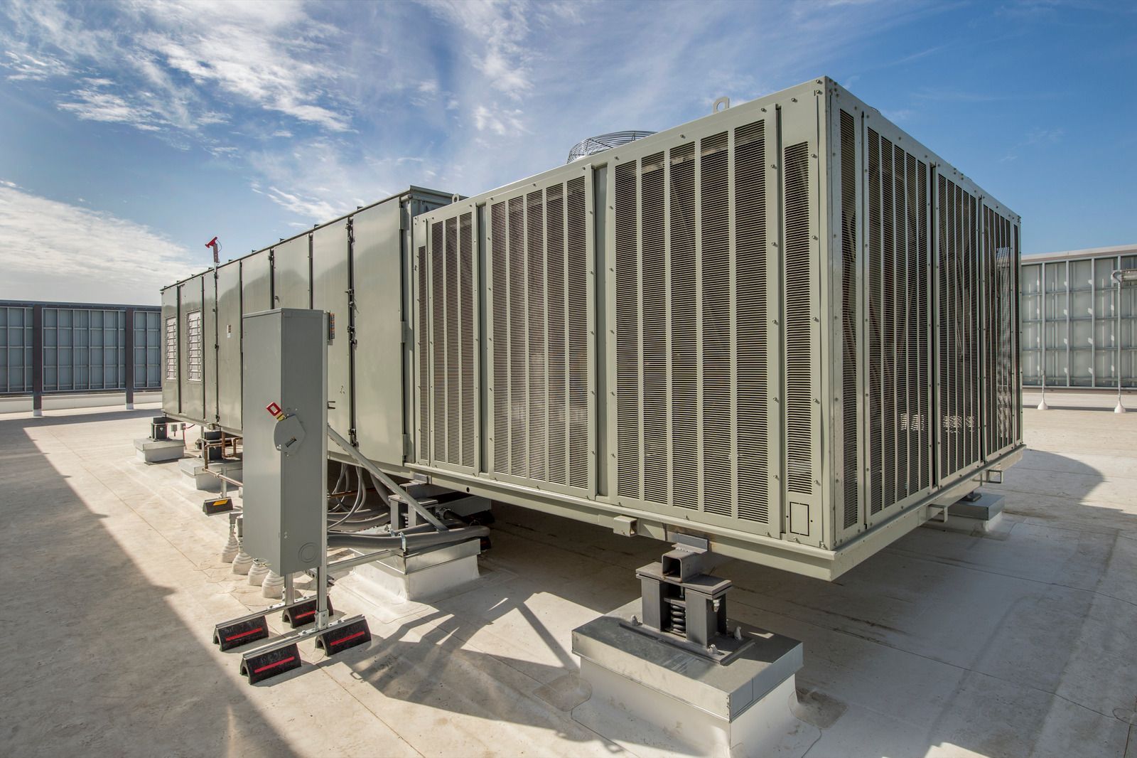 Large HVAC unit on a rooftop, gray metal with protective grates, supported by concrete blocks and metal framework.