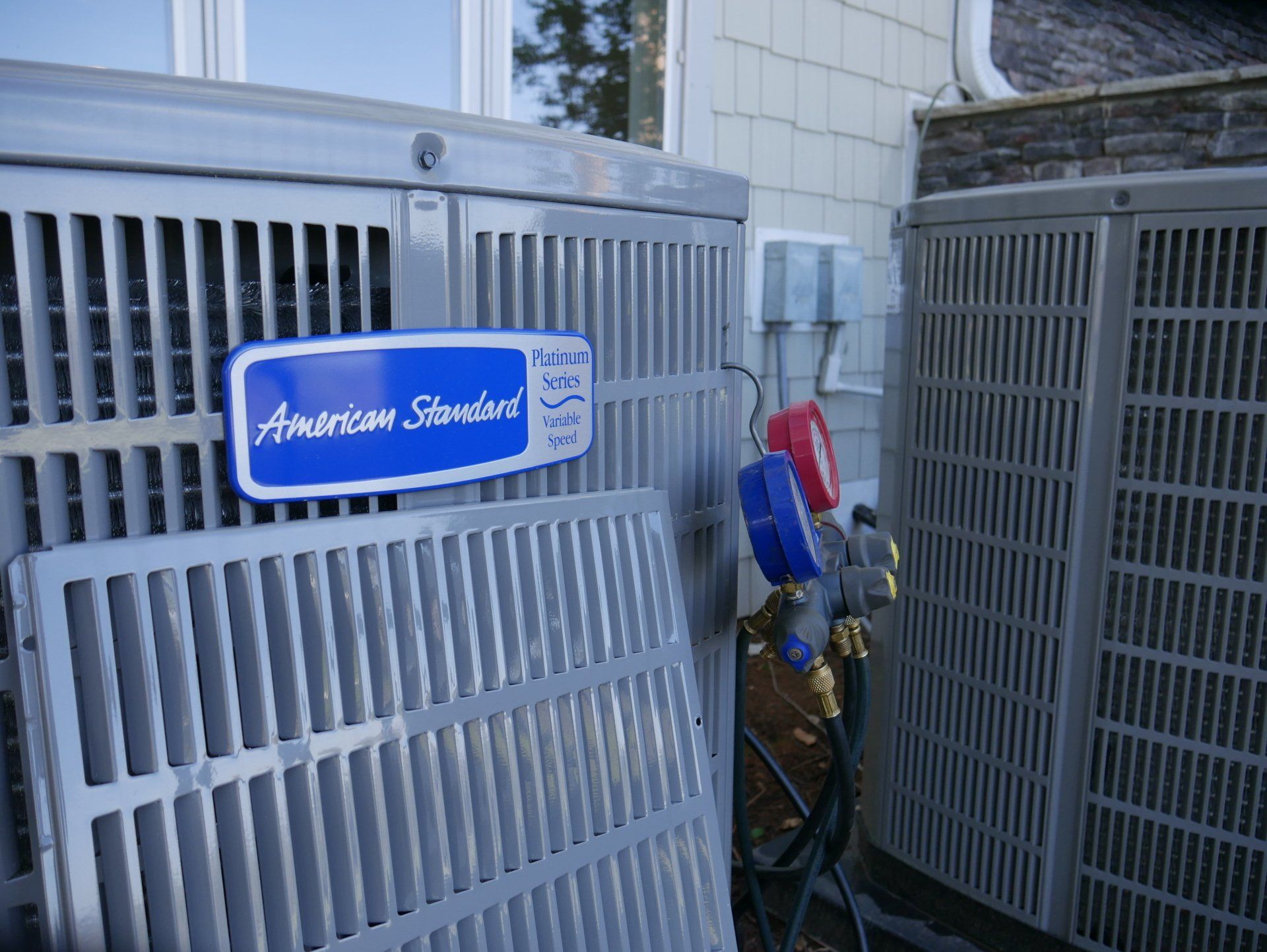 Two American Standard air conditioning units being serviced, with gauges attached. One unit has a removed grate.