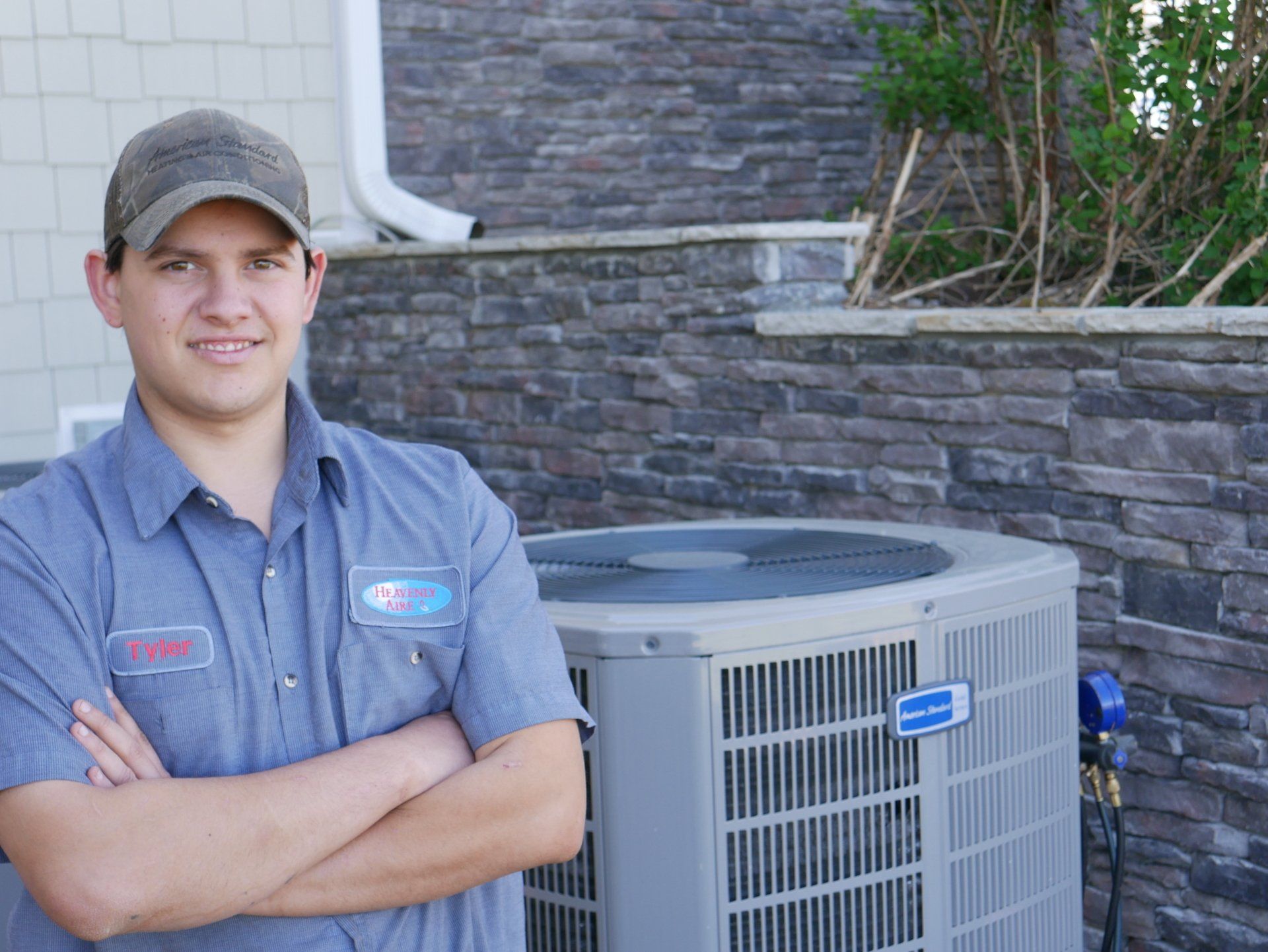 HVAC technician in blue work shirt and cap standing next to an air conditioning unit, arms crossed.