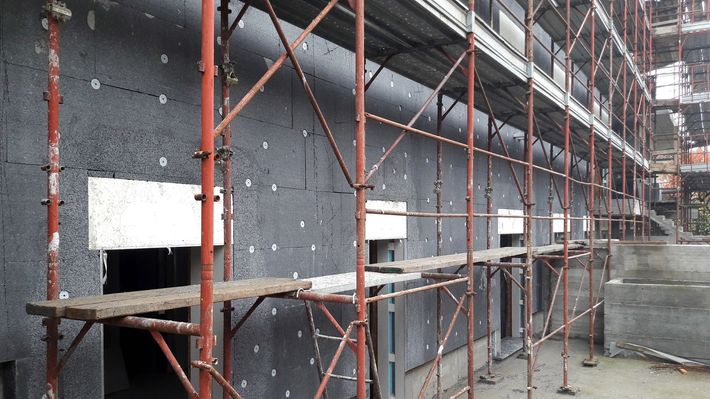 Facciata di un edificio con impalcature, pareti grigie e supporti metallici rossi, in un cantiere edile.