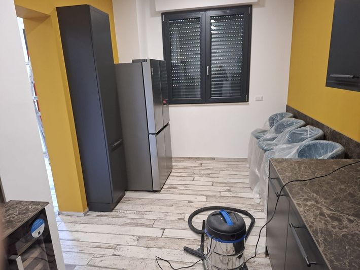 Cucina moderna con mobili grigi, ripiani scuri, pavimento piastrellato bianco, finestra e aspirapolvere sul pavimento.