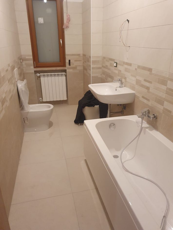 Piccolo bagno con vasca, lavandino, WC, finestra e pareti rivestite con piastrelle beige.