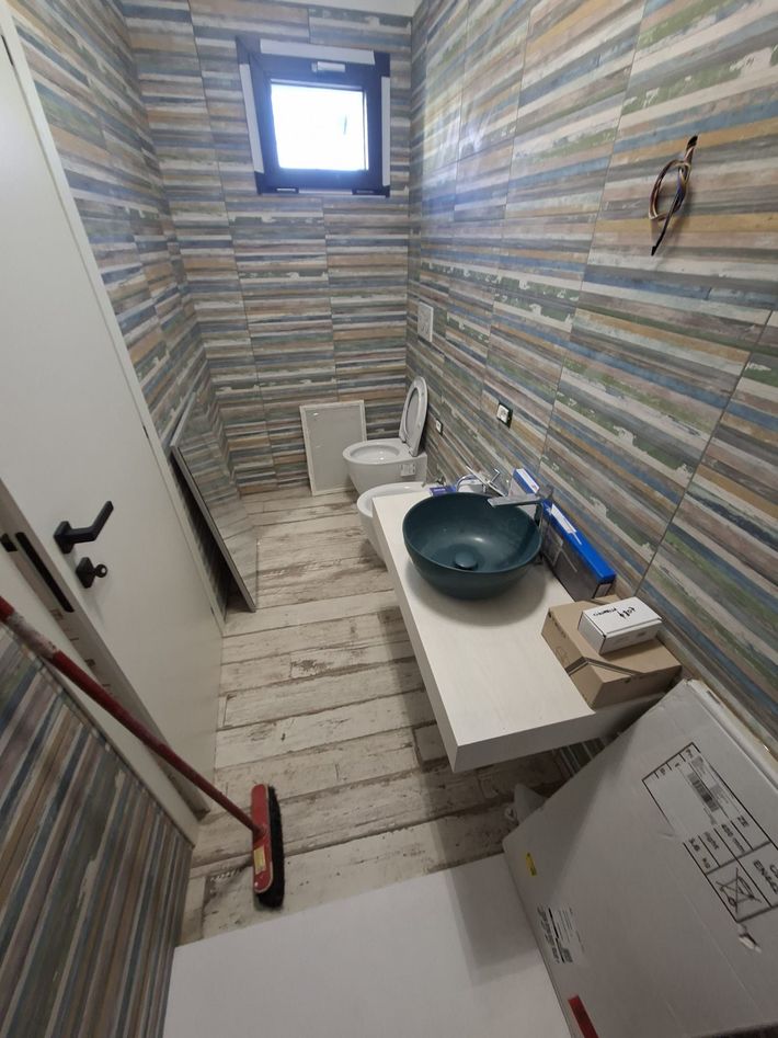 Bagno stretto piastrellato con WC, lavandino, finestra e accessori per la pulizia.