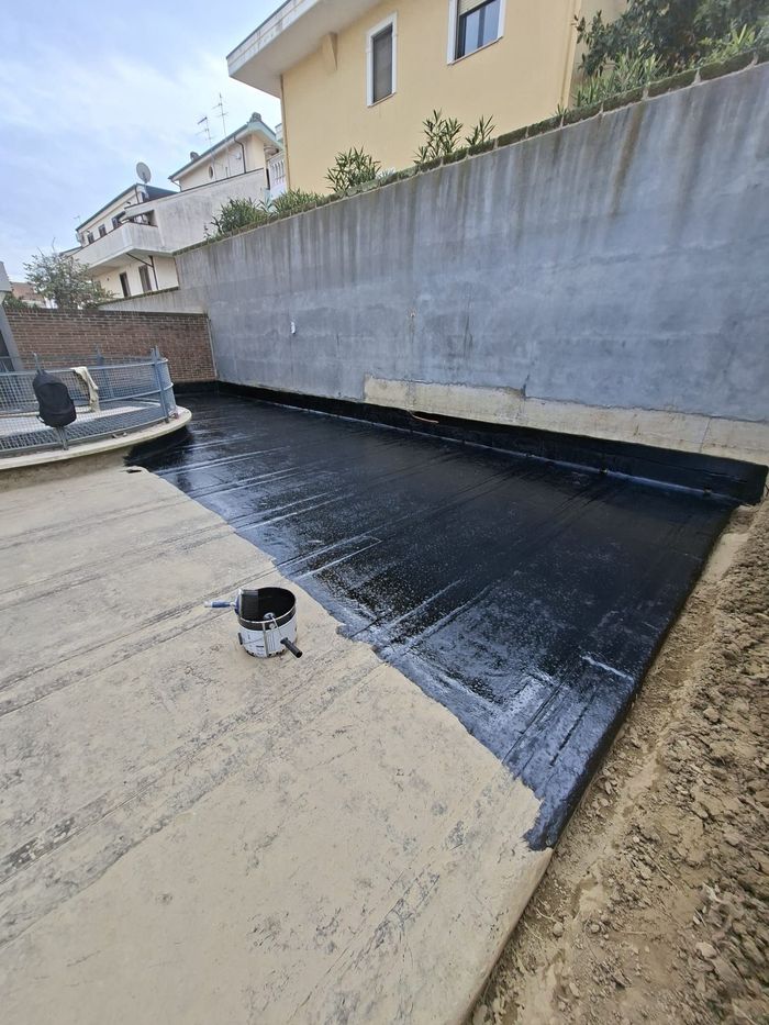 Applicazione di una membrana impermeabile nera su una giunzione di una parete del tetto, accanto a una casa beige.