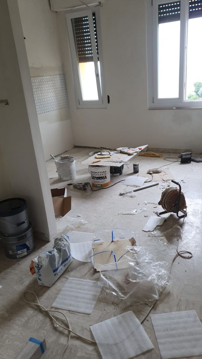 Stanza disordinata con detriti, carte e oggetti rotti sparsi sul pavimento vicino a due finestre.