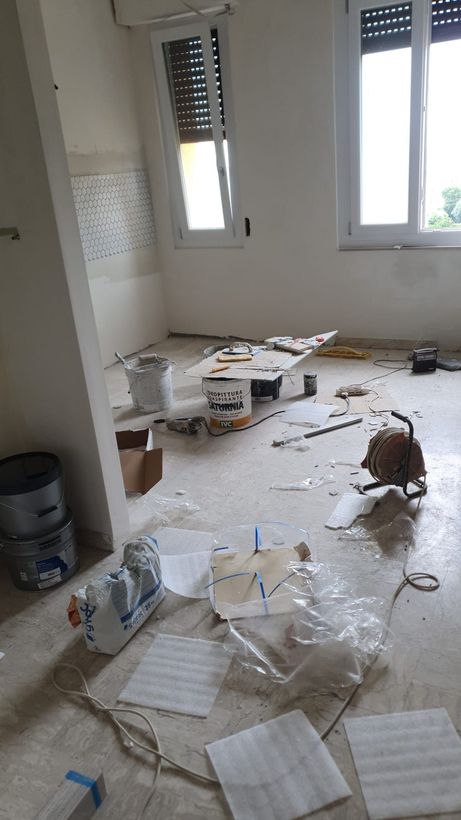 Stanza disordinata con detriti, carte e oggetti rotti sparsi sul pavimento vicino a due finestre.