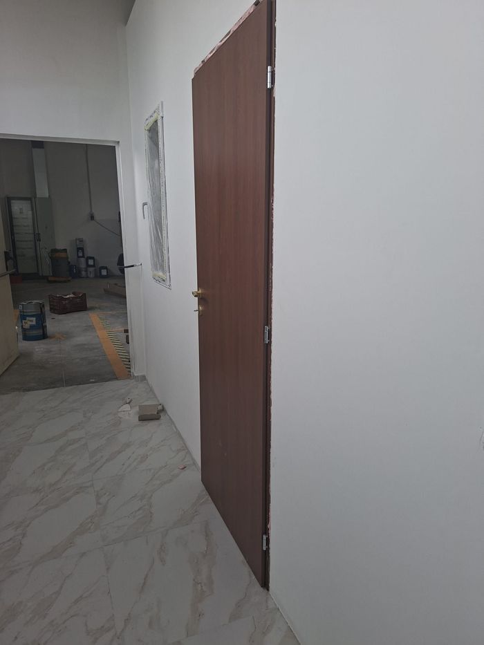 Stretto corridoio con pareti bianche, una porta marrone e una stanza non finita visibile in fondo.