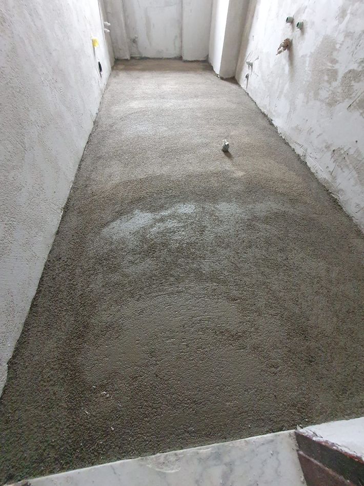 Stretto corridoio in cemento grezzo con pareti bianche e pavimento grigio ruvido.