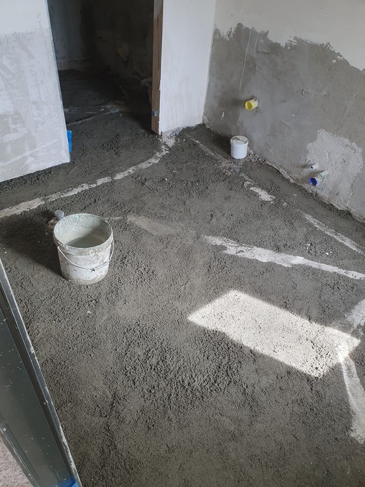 Stanza non finita con pavimento in cemento bagnato, secchi bianchi e pareti intonacate di grigio vicino a una porta.