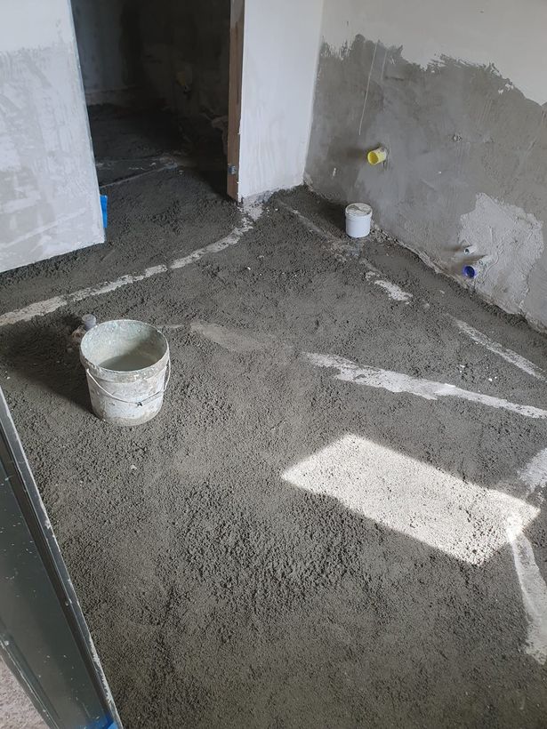 Stanza non finita con pavimento in cemento bagnato, secchi bianchi e pareti intonacate di grigio vicino a una porta.