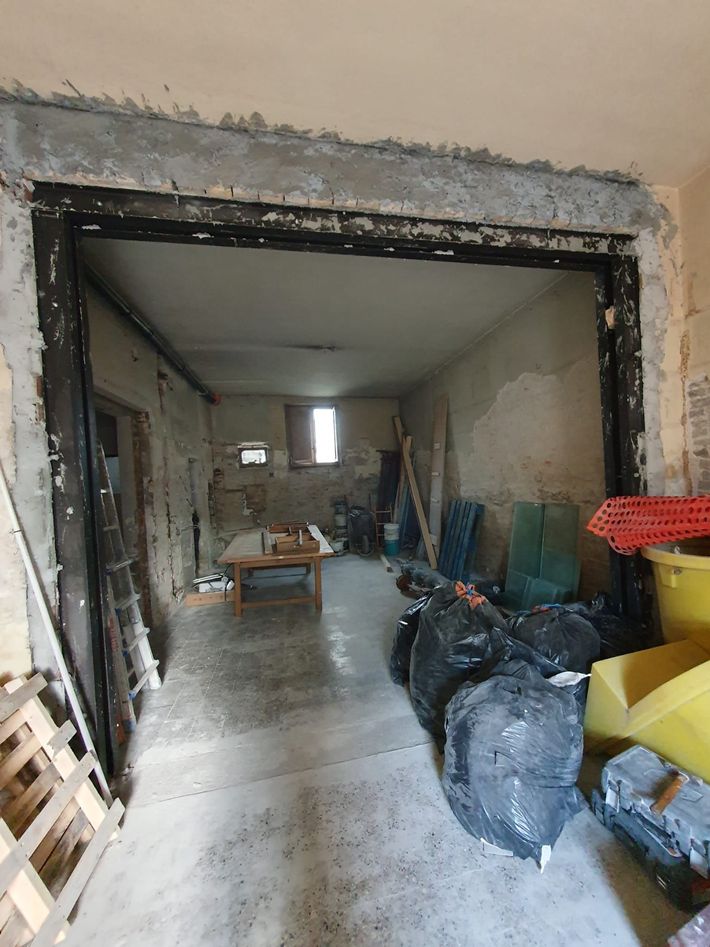 Stanza ristrutturata con porta non rifinita, attrezzi, sacchi per detriti e materiali accatastati lungo le pareti.