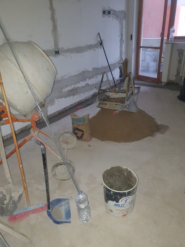 Locale adibito a cantiere con pareti intonacate, attrezzi, secchi e terra sparsa sul pavimento.