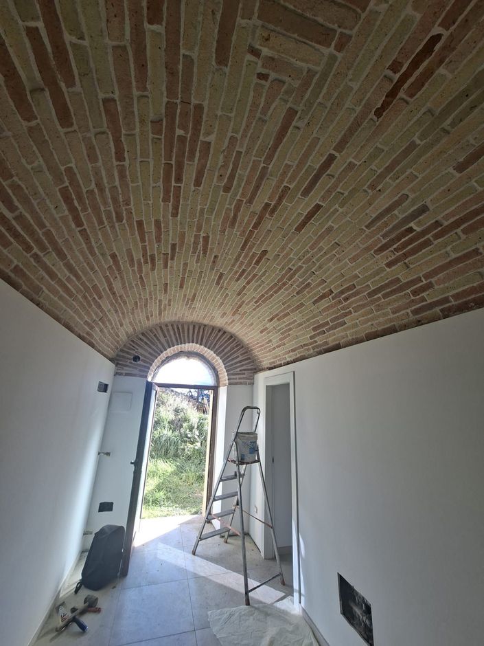 Stretto corridoio con soffitto a volta a botte in mattoni, pareti bianche, scala e luce proveniente da una porta ad arco.